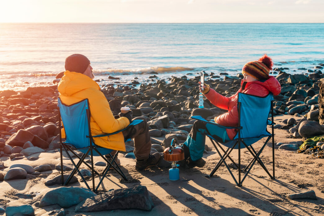 Two people on beach chairs together on winter beach, making hot tea, coffee on sunny blue sky day and the sea in background. British cold winter. Lifestyle tourism concept - Gringo Gazette