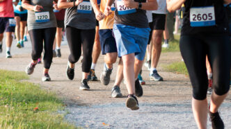 Group of runners in a race, visible bib numbers, midstride on a gravel path in a park.