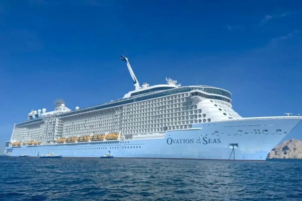Large white cruise ship Ovation of the Seas anchored on calm blue ocean under a clear sky, with multiple decks and balconies visible.