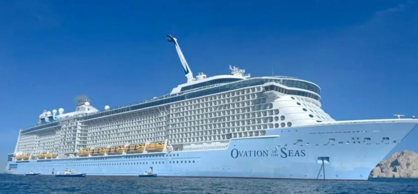 Large white cruise ship Ovation of the Seas anchored on calm blue ocean under a clear sky, with multiple decks and balconies visible.