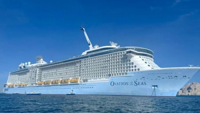 Large white cruise ship Ovation of the Seas anchored on calm blue ocean under a clear sky, with multiple decks and balconies visible.