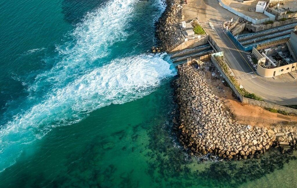 Aerial view of turquoise sea meeting a rocky breakwater with a road and harbor structures along the shore.