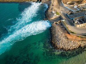 Aerial view of turquoise sea meeting a rocky breakwater with a road and harbor structures along the shore.
