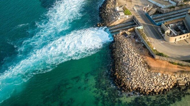 Aerial view of turquoise sea meeting a rocky breakwater with a road and harbor structures along the shore.