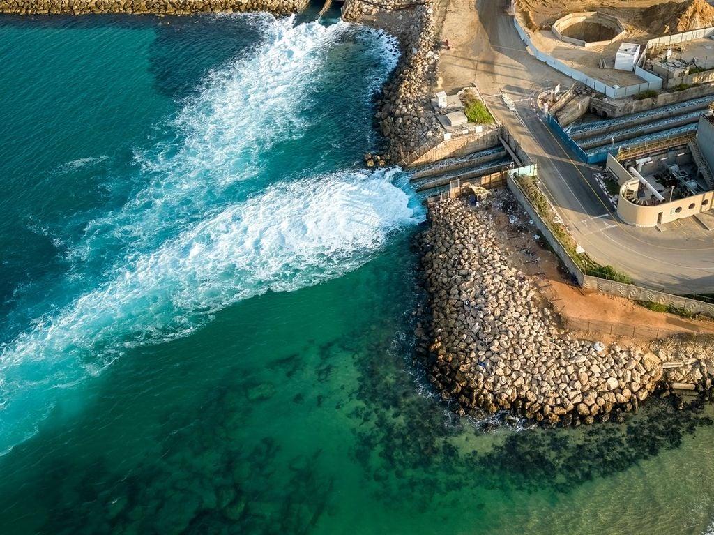 Aerial view of turquoise sea meeting a rocky breakwater with a road and harbor structures along the shore.