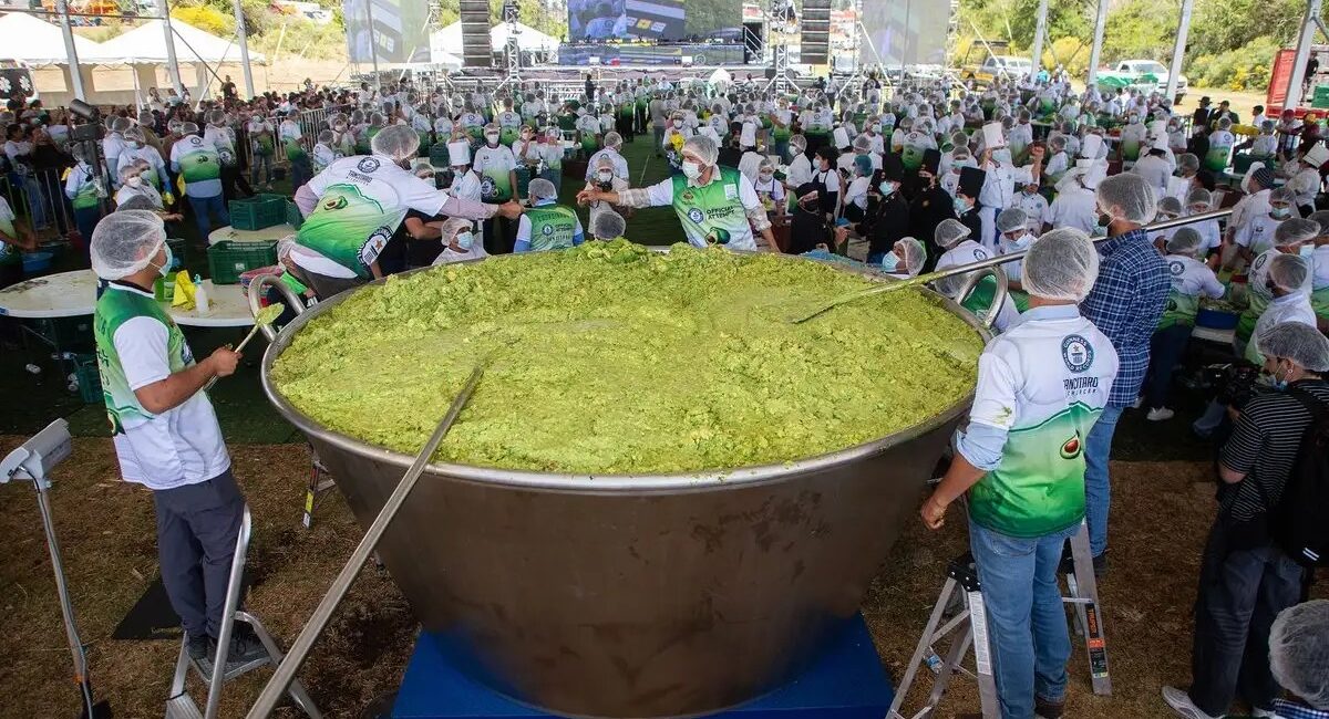 Large stainless steel cauldron filled with bright green food, volunteers in green-and-white vests stirring it under a festival tent.