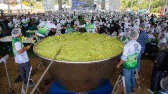 Large stainless steel cauldron filled with bright green food, volunteers in green-and-white vests stirring it under a festival tent.