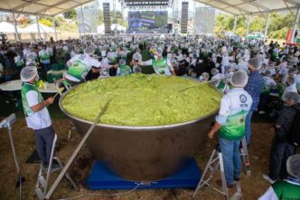 Large stainless steel cauldron filled with bright green food, volunteers in green-and-white vests stirring it under a festival tent.