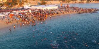 Crowded beach event with swimmers in clear blue water and tents along the shore.