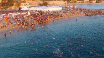 Crowded beach event with swimmers in clear blue water and tents along the shore.