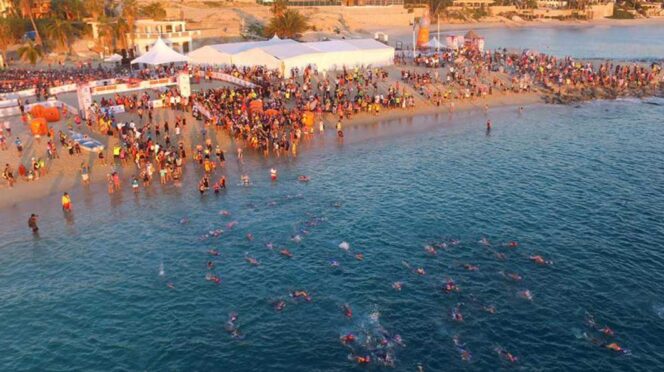 Crowded beach event with swimmers in clear blue water and tents along the shore.