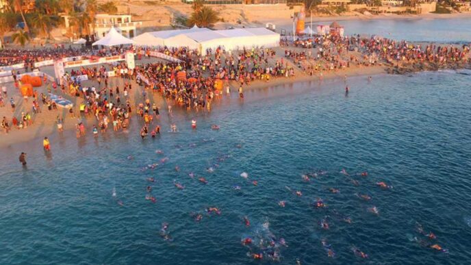 Crowded beach event with swimmers in clear blue water and tents along the shore.