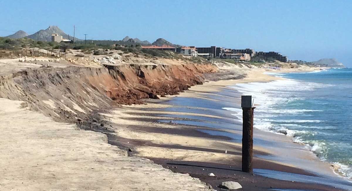 Coastal beach with eroded brown cliff, sandy shore, and calm blue sea; distant buildings on the coastline.