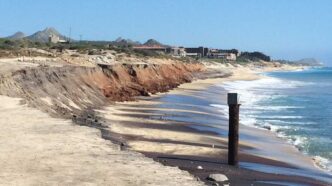 Coastal beach with eroded brown cliff, sandy shore, and calm blue sea; distant buildings on the coastline.