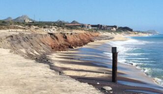 Coastal beach with eroded brown cliff, sandy shore, and calm blue sea; distant buildings on the coastline.