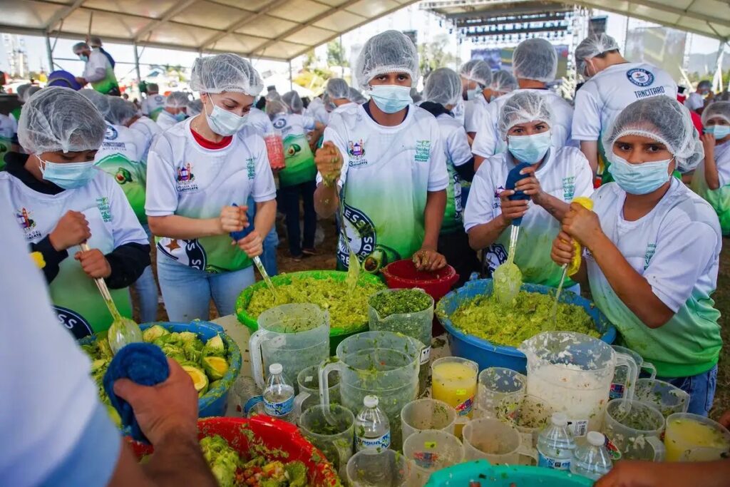 Group of young volunteers in hairnets and masks stirring large bowls of green salsa under a canopy at a community food event.