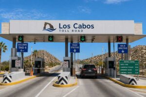 Toll booth lane at Los Cabos Toll Road with green downward arrows and a black SUV approaching the gate, desert hills in the background.