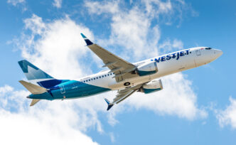 WestJet airliner in blue and white livery flying through a bright, partly cloudy sky.