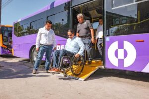 Wheelchair user being assisted off a purple bus with a fold-out ramp as two men help nearby, showing accessible transit.