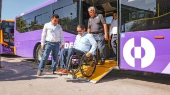 Wheelchair user being assisted off a purple bus with a fold-out ramp as two men help nearby, showing accessible transit.