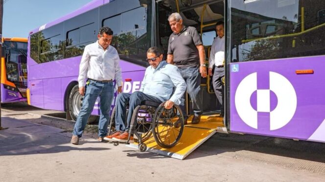Wheelchair user being assisted off a purple bus with a fold-out ramp as two men help nearby, showing accessible transit.