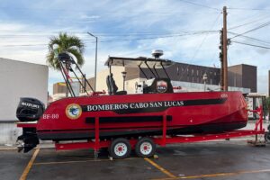 Red firefighting boat on a trailer, labeled Bomberos Cabo San Lucas, with Suzuki outboard motor and equipment on top, parked in a lot.