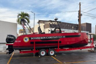Red firefighting boat on a trailer, labeled Bomberos Cabo San Lucas, with Suzuki outboard motor and equipment on top, parked in a lot.