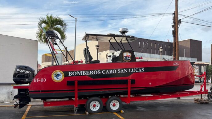 Red firefighting boat on a trailer, labeled Bomberos Cabo San Lucas, with Suzuki outboard motor and equipment on top, parked in a lot.