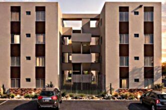 Beige apartment complex with two four-story towers and a central open walkway; gated entrance and parked cars in front.