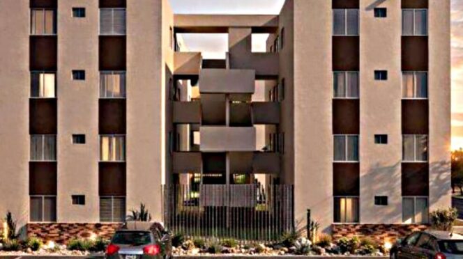Beige apartment complex with two four-story towers and a central open walkway; gated entrance and parked cars in front.