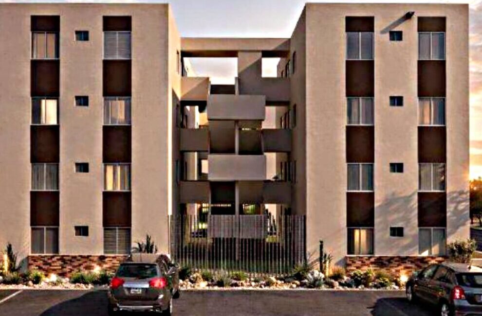 Beige apartment complex with two four-story towers and a central open walkway; gated entrance and parked cars in front.