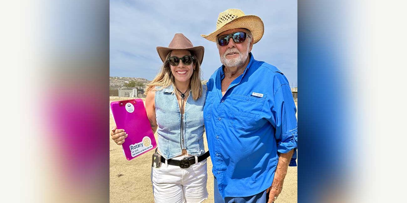 Smiling woman in a brown hat and denim vest stands beside an older man in a blue shirt and straw hat outdoors; she holds a pink Rotary clipboard.