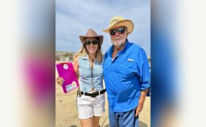 Smiling woman in a brown hat and denim vest stands beside an older man in a blue shirt and straw hat outdoors; she holds a pink Rotary clipboard.