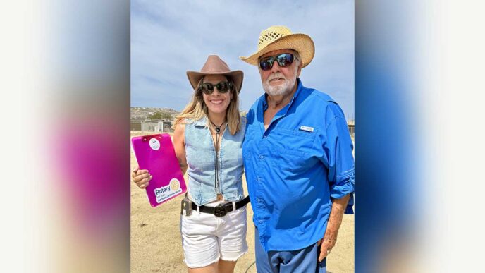 Smiling woman in a brown hat and denim vest stands beside an older man in a blue shirt and straw hat outdoors; she holds a pink Rotary clipboard.