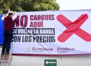 Two men on a ladder hang a large protest banner with a bold red X, calling out high prices; government logos shown at the bottom.