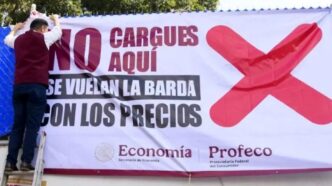 Two men on a ladder hang a large protest banner with a bold red X, calling out high prices; government logos shown at the bottom.