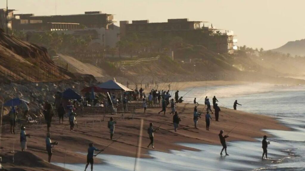 Crowded beach at sunset with many people fishing along the shoreline and tents set up nearby. Buildings line the distant coast.