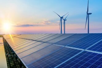 Solar panels in the foreground with wind turbines on the horizon at sunset, representing renewable energy.