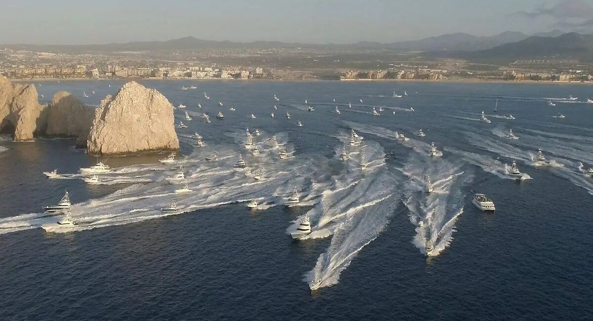 Aerial view of a harbor with numerous white speedboats creating wakes near rocky islets and a distant city shoreline.