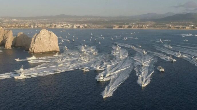 Aerial view of a harbor with numerous white speedboats creating wakes near rocky islets and a distant city shoreline.