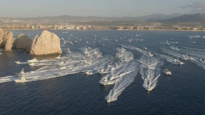 Aerial view of a harbor with numerous white speedboats creating wakes near rocky islets and a distant city shoreline.