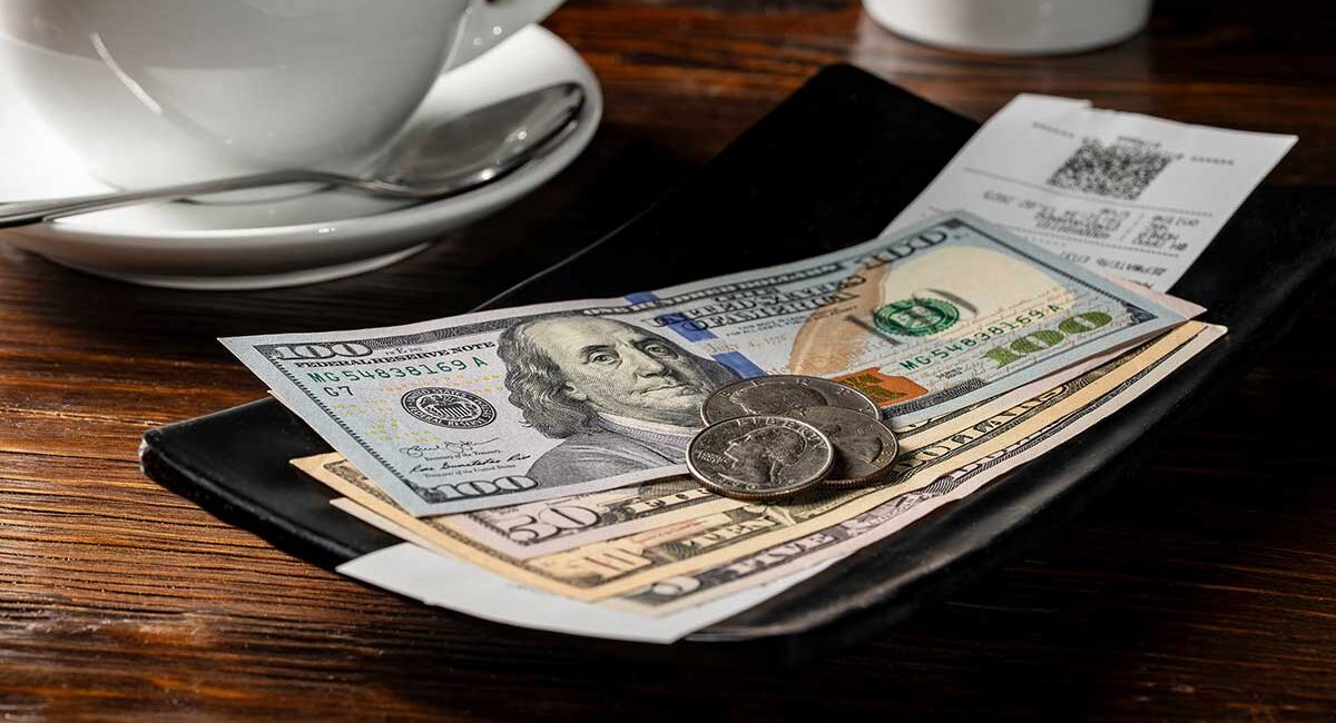 Open wallet on a wooden table with $100 bills and coins, beside a coffee cup and a receipt behind it.