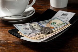 Open wallet on a wooden table with $100 bills and coins, beside a coffee cup and a receipt behind it.