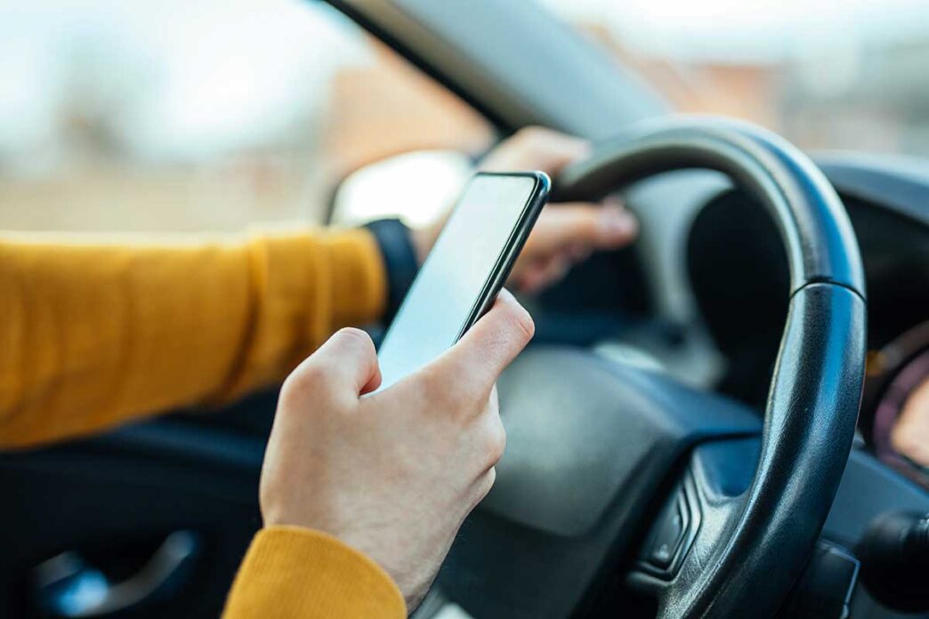 Driver in a mustard-colored sweater holds a smartphone while gripping the steering wheel in a car.