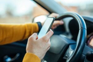 Driver in a mustard-colored sweater holds a smartphone while gripping the steering wheel in a car.