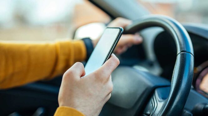 Driver in a mustard-colored sweater holds a smartphone while gripping the steering wheel in a car.