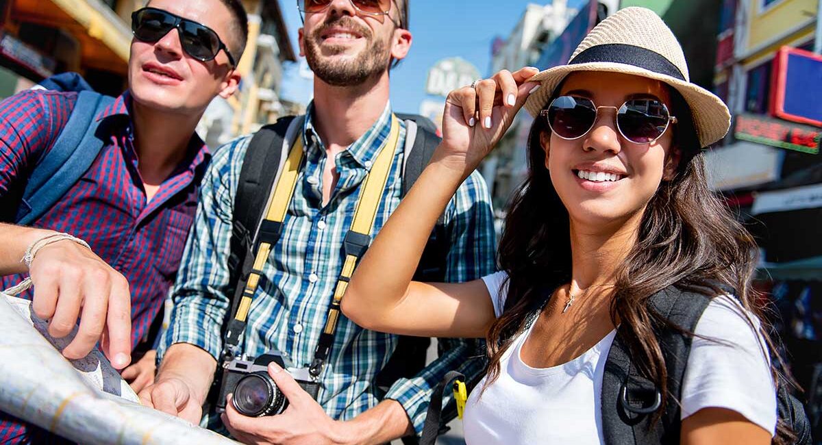 Three friends posing for a photo on a sunny city street; one holds a camera and wears a backpack, the others wear sunglasses and hats, smiling at the camera.