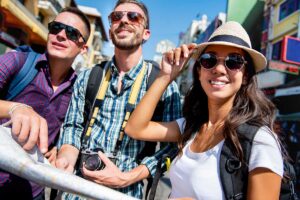 Three friends posing for a photo on a sunny city street; one holds a camera and wears a backpack, the others wear sunglasses and hats, smiling at the camera.
