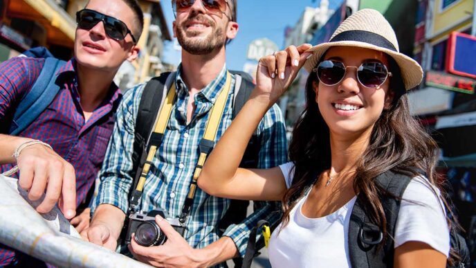 Three friends posing for a photo on a sunny city street; one holds a camera and wears a backpack, the others wear sunglasses and hats, smiling at the camera.