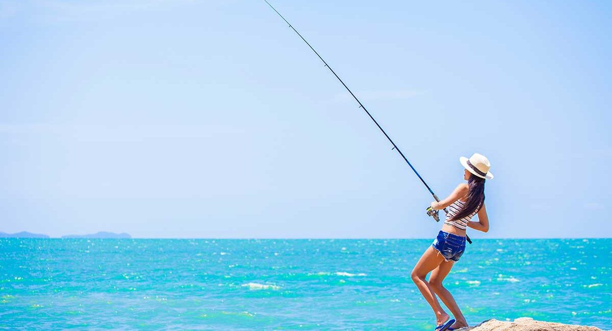 Woman standing on a rocky beach, fishing rod cast over turquoise ocean under a clear blue sky, wearing a sun hat and shorts.